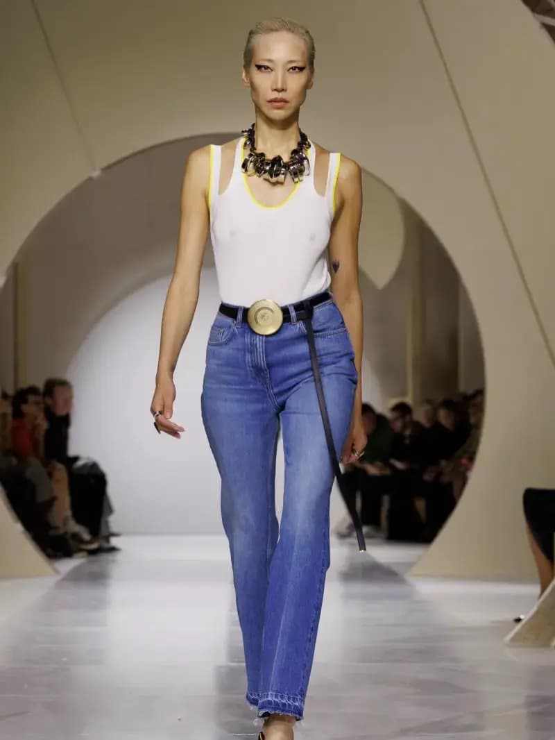 Runway model in a white tank, gold statement necklace, flared blue jeans and the compact belt with a long trailing strap