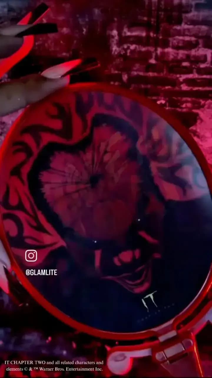 Hand holding a round red mirror printed with a dark Pennywise face and IT logo against a brick wall