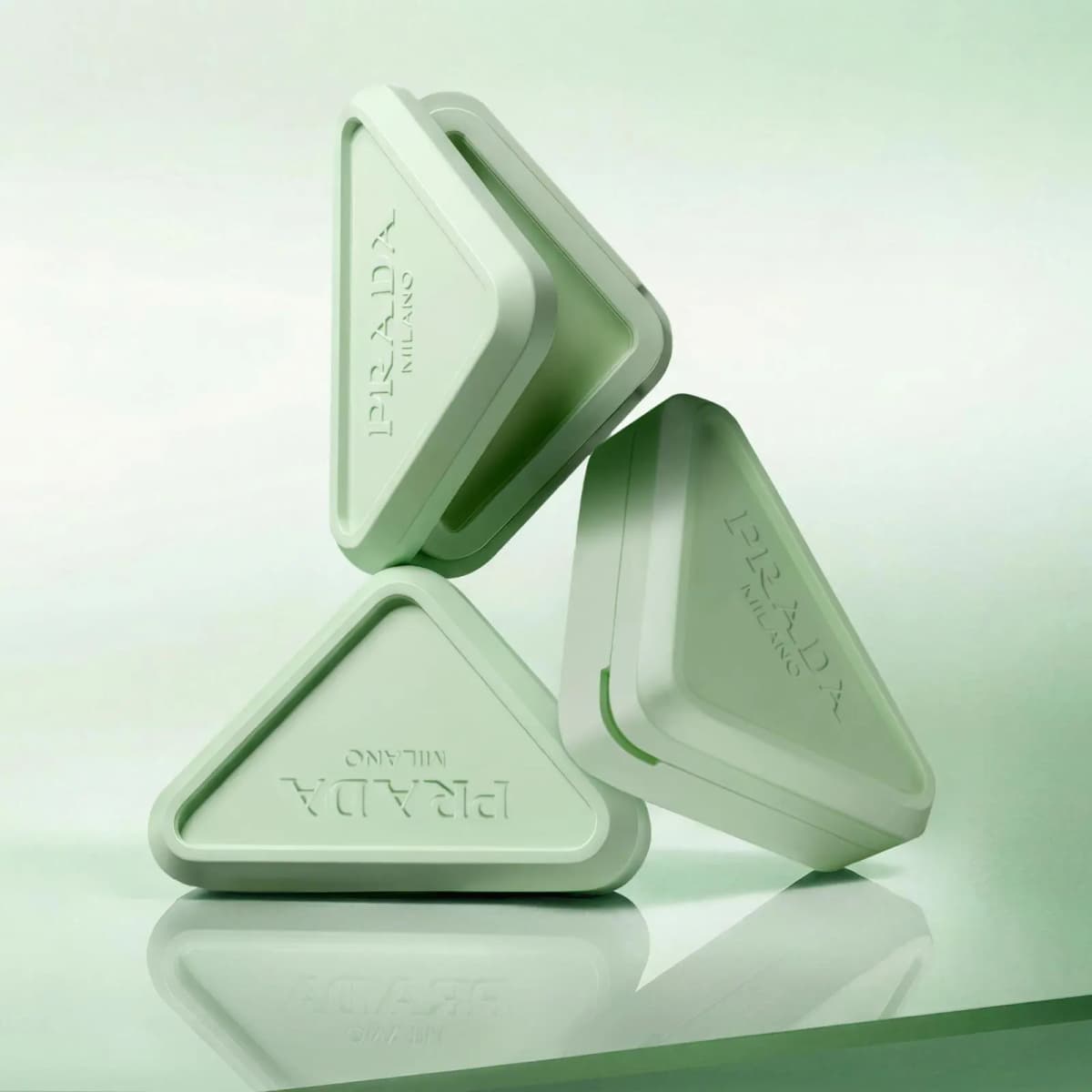 Three closed mint-green Prada Milano triangular compacts stacked and balanced on a reflective green surface