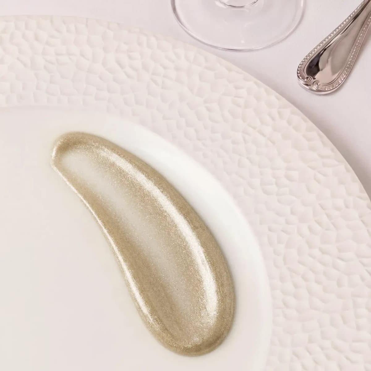 Swatch of shimmering pale gold highlighter on a textured white plate beside a wine glass and silver spoon