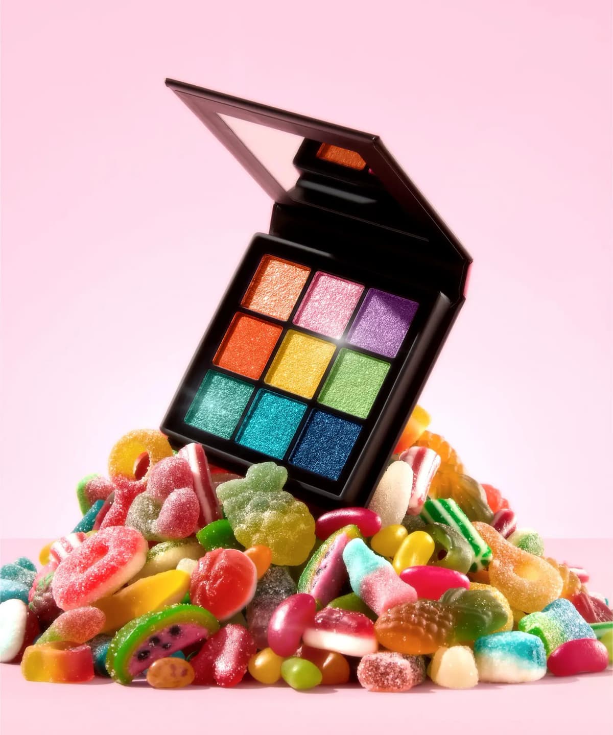 Open rainbow shimmer palette perched on a pile of colourful sugar-coated gummy sweets against a pink background