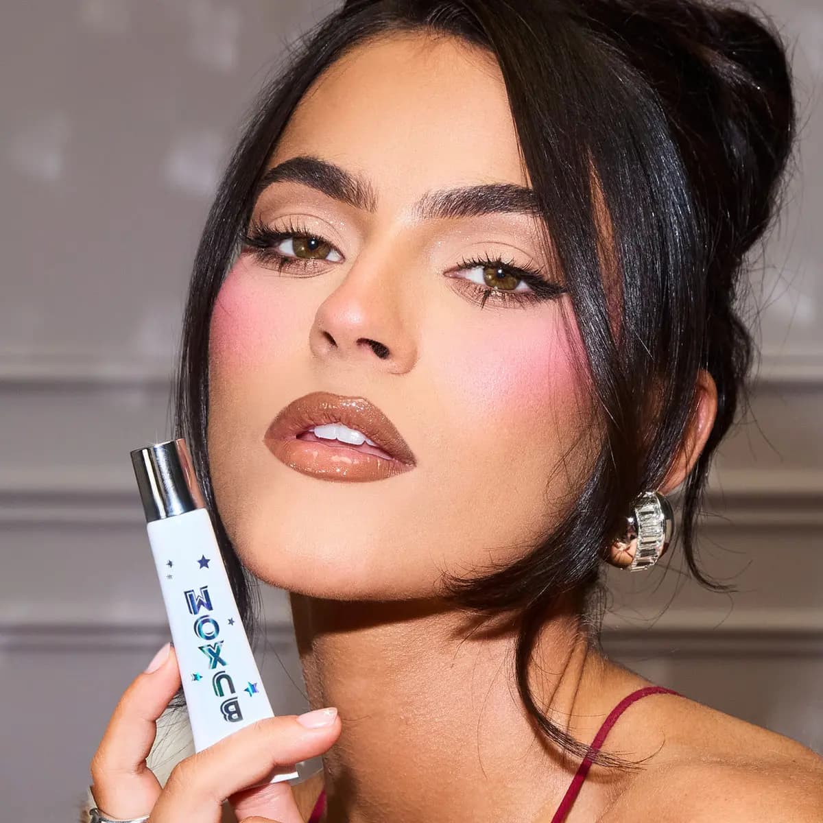 Model with dark hair and glossy nude-shimmer lips holding the white BUXOM Last Call tube