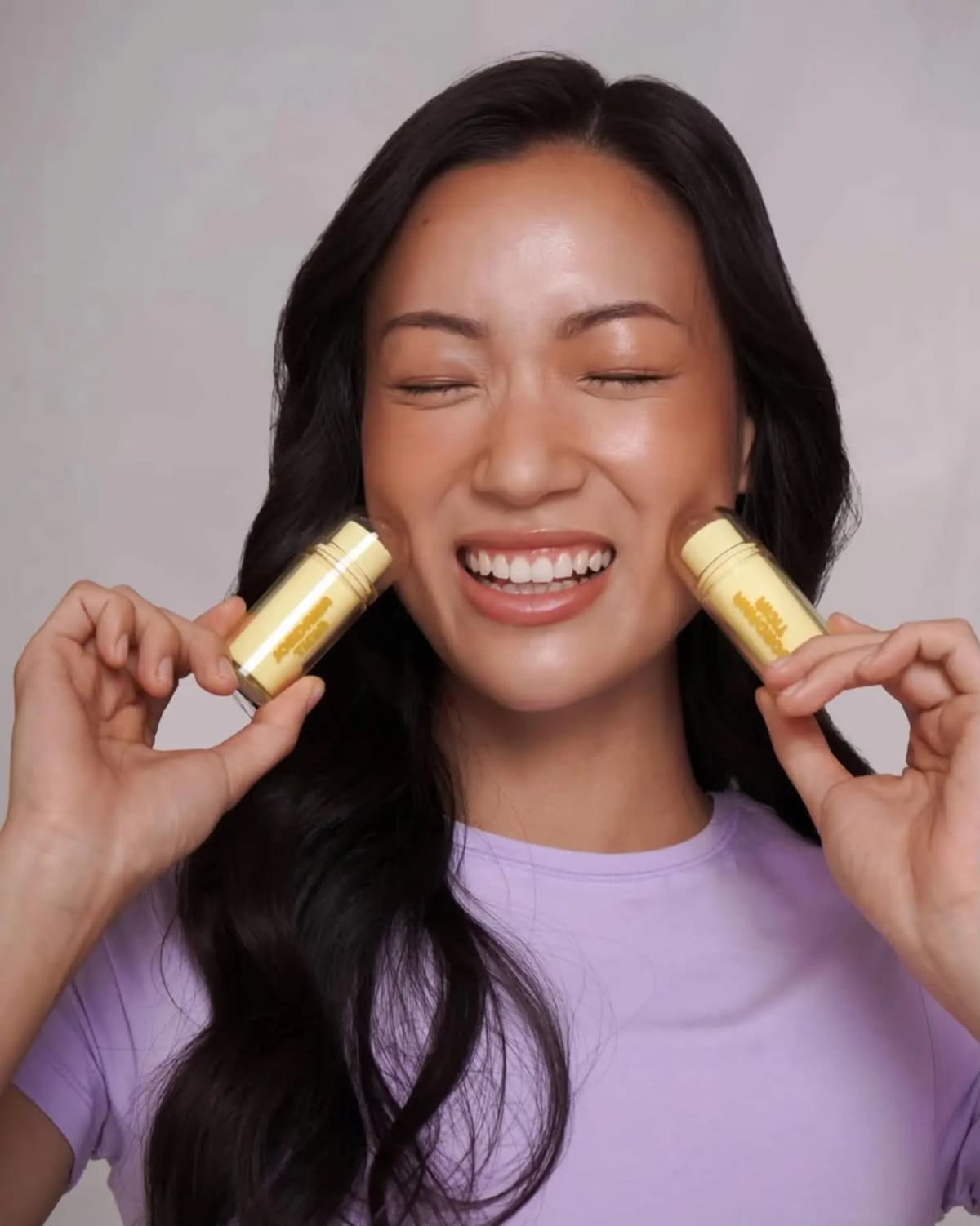 Smiling model in a lilac T-shirt presses two yellow bronzer sticks to her cheeks, eyes closed