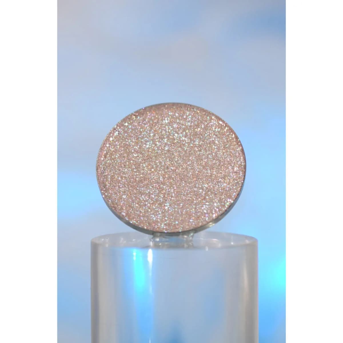 Sparkly pinkish-silver pressed pan with holographic glitter balanced on a clear acrylic block against a blue backdrop
