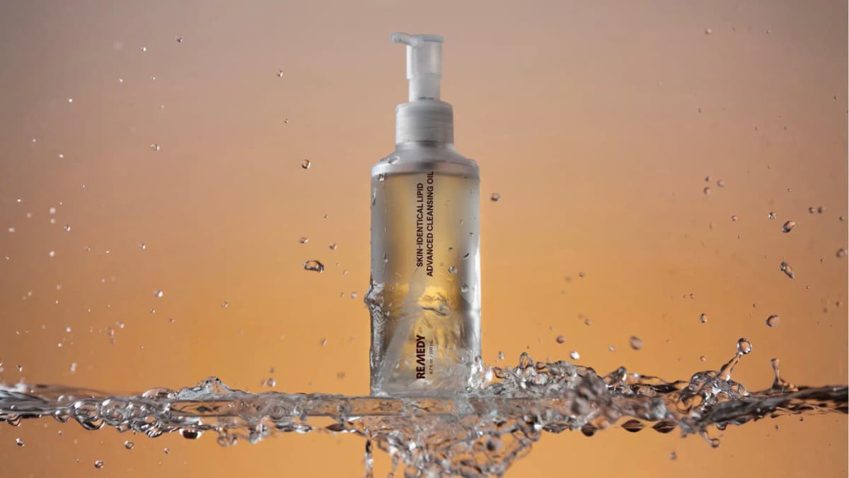 Frosted bottle of golden cleansing oil rising from a splash of water against a warm orange backdrop
