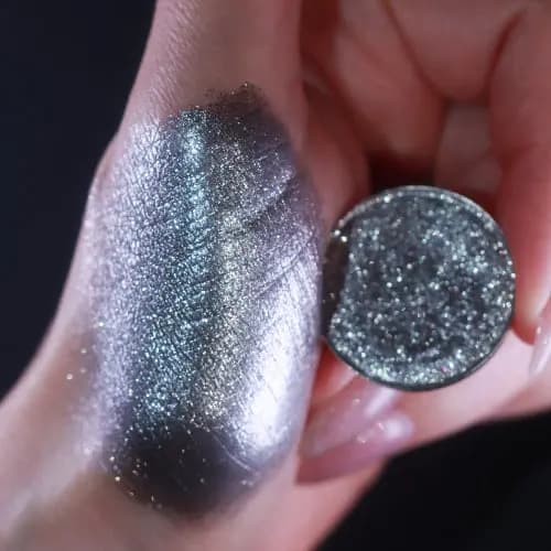 Hand holds a sparkly gunmetal shimmer shadow pan beside a reflective silver swatch shifting from lilac to blue-green.