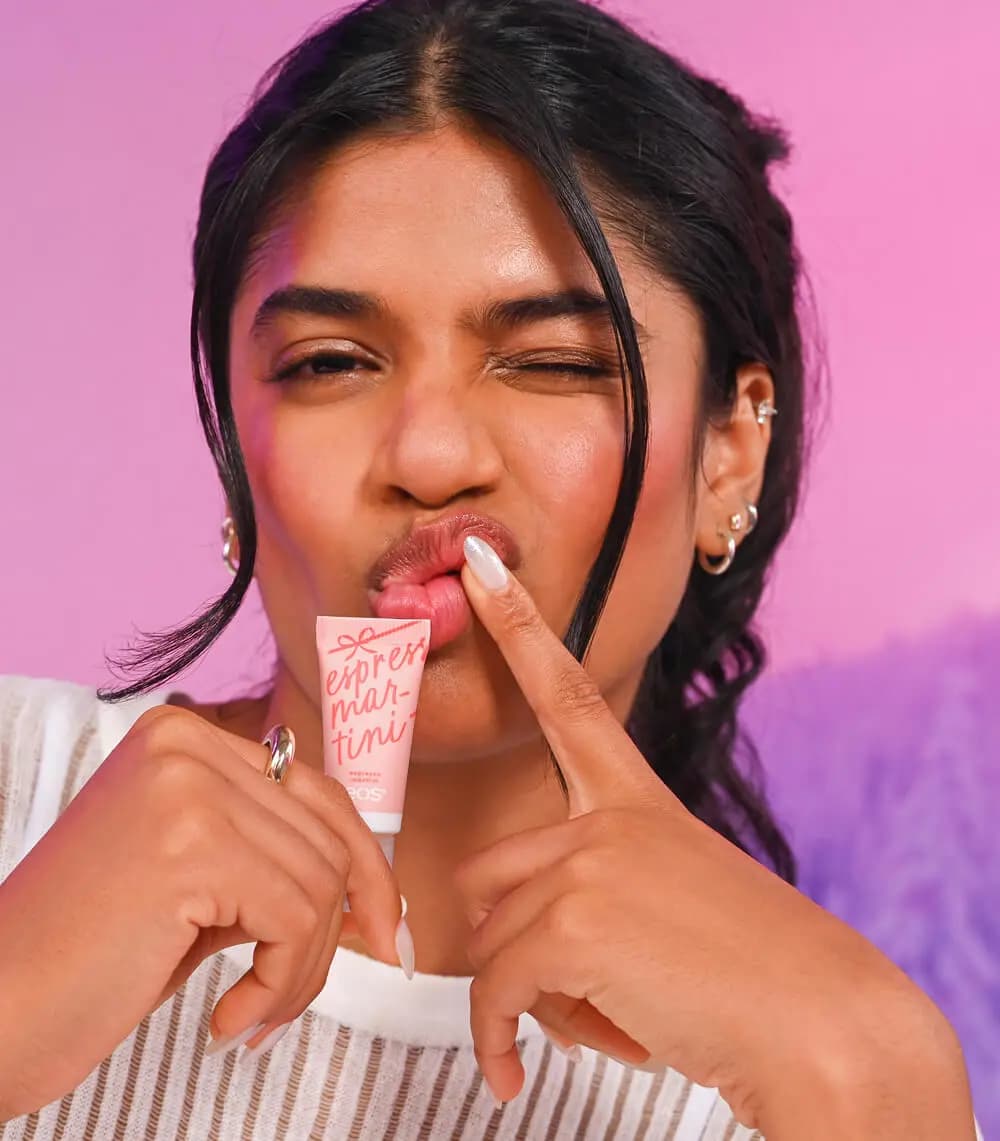 Winking model with glossy pink lips holds a peach eos Espresso Martini mini balm tube against a pink and purple background