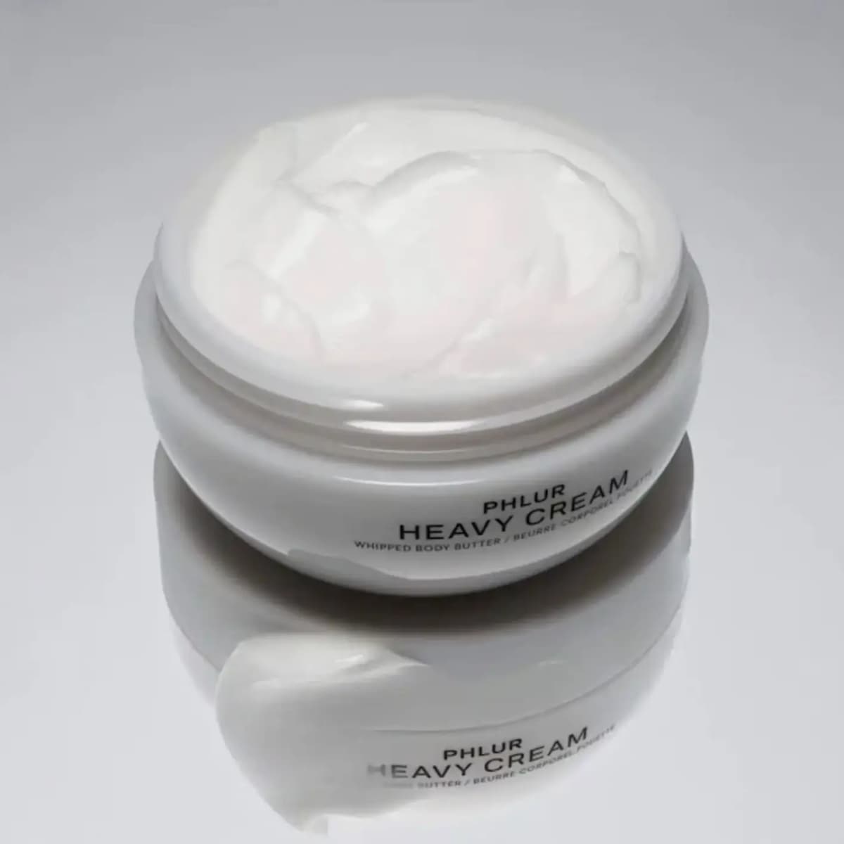 Open PHLUR Heavy Cream jar showing fluffy white whipped body butter, with a swatch on a glossy grey reflective surface