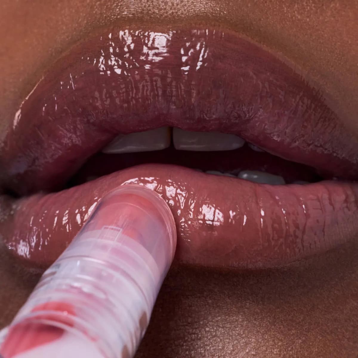 Clear applicator tip pressing a glossy rosy-brown tint onto the lower lip of a model with deep skin