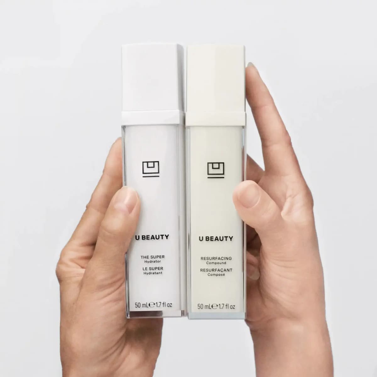 Two hands holding 50 mL bottles of The SUPER Hydrator and Resurfacing Compound against a pale grey background