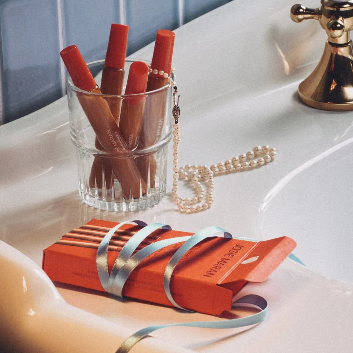 Four capped fragrance mist tubes in a glass cup with pearls, next to a red ribbon-wrapped box on a bathtub edge.