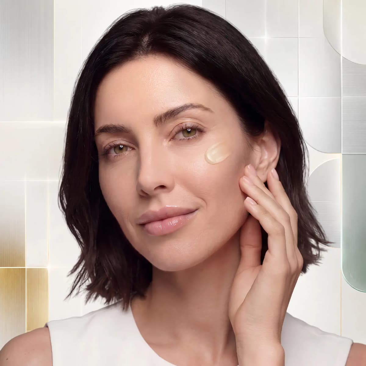 Model with a bob cupping her jaw, a swipe of glossy golden oil on her cheekbone, against metallic tiles