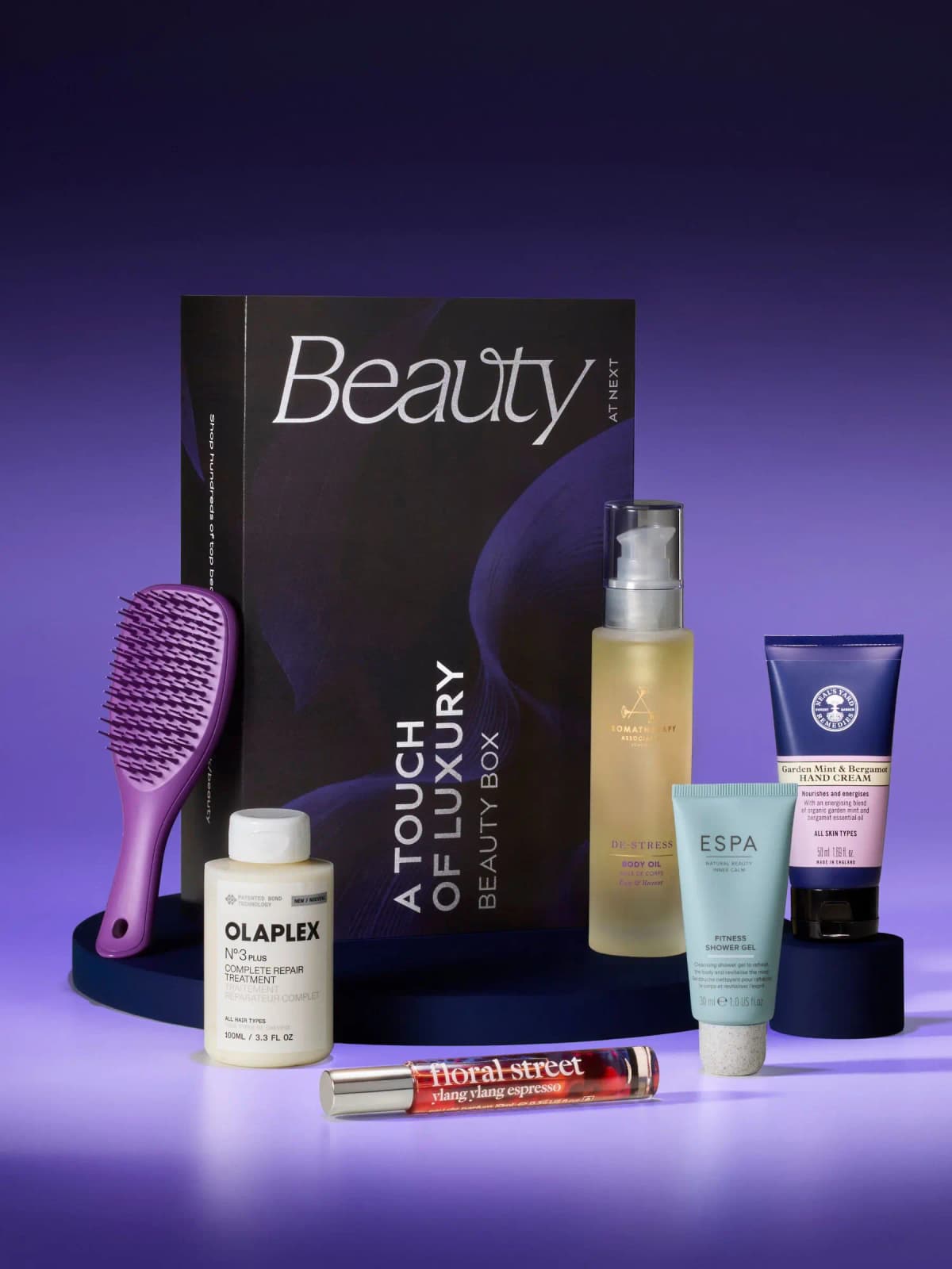 Next Beauty A Touch of Luxury box with Olaplex, body oil, ESPA gel, hand cream, purple brush and perfume on purple backdrop