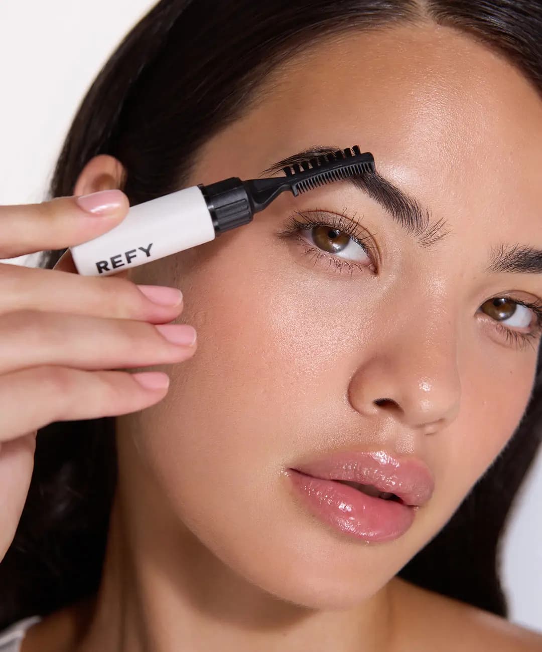 Model combing their brow upward with a white REFY brow gel wand, lips glossy pink