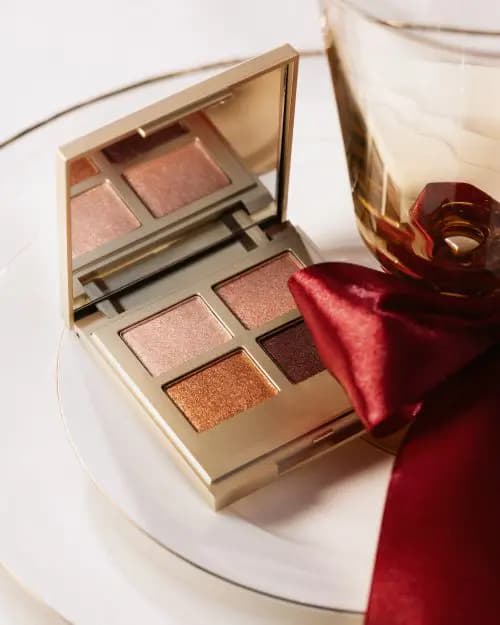 Gold compact eyeshadow quad in shimmering rose, copper and plum shades next to a glass tied with a red ribbon