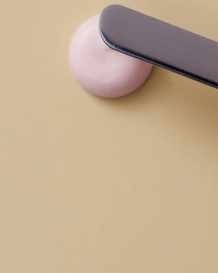 Metal spatula pressing into a small dollop of pink primer cream on a tan surface