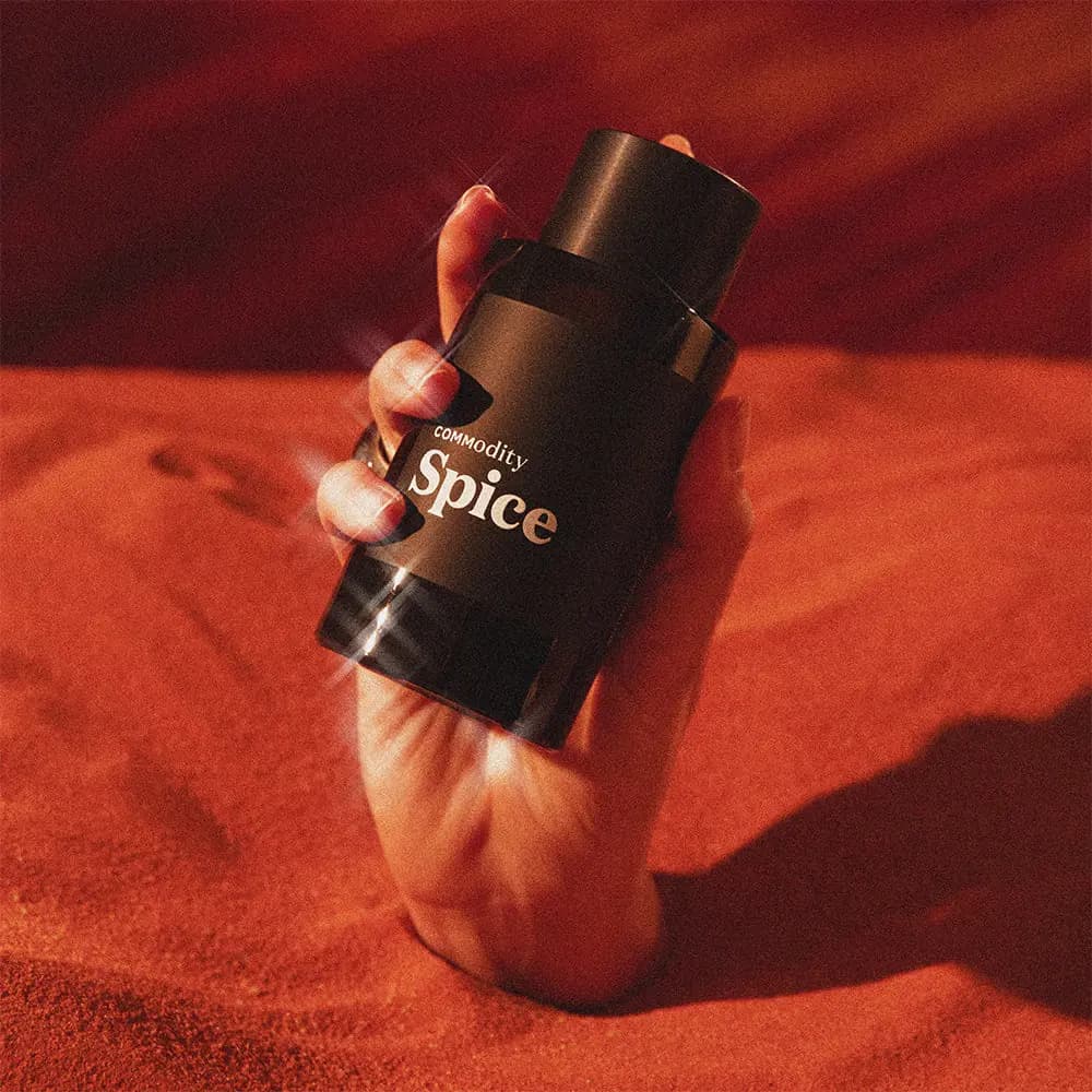 Hand rising from red-orange sand, gripping a black Commodity Spice bottle with sparkling highlights