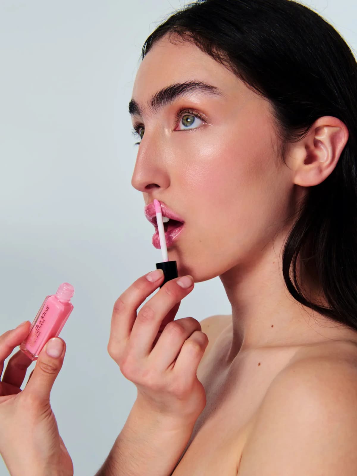 Profile of a model applying pink lip oil with a wand while holding the open Les Filles en Rouje bottle