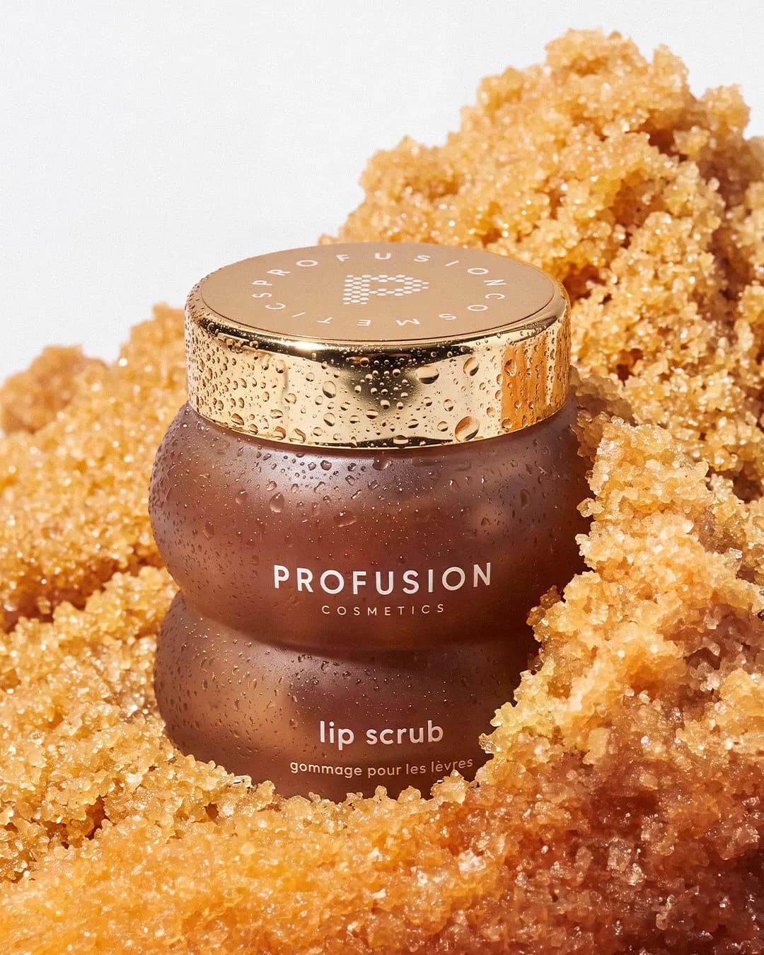 Profusion lip scrub jar with a gold lid, beaded with water droplets and nestled in golden sugar crystals.