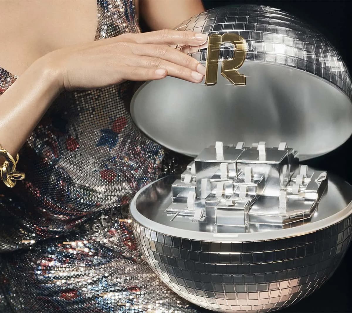 Model in shimmering metallic dress opens a mirrored disco-ball calendar with gold Rabanne R, revealing silver numbered