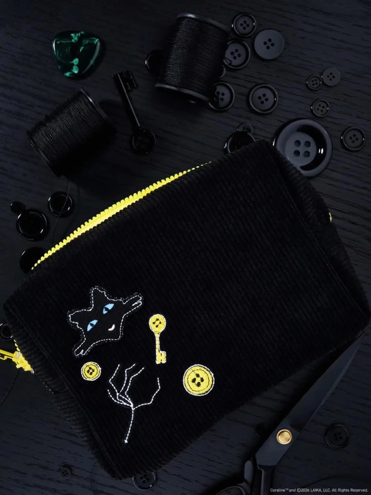 Black corduroy zip pouch with yellow zipper, embroidered black cat, key and button motifs, on dark wood with thread spools