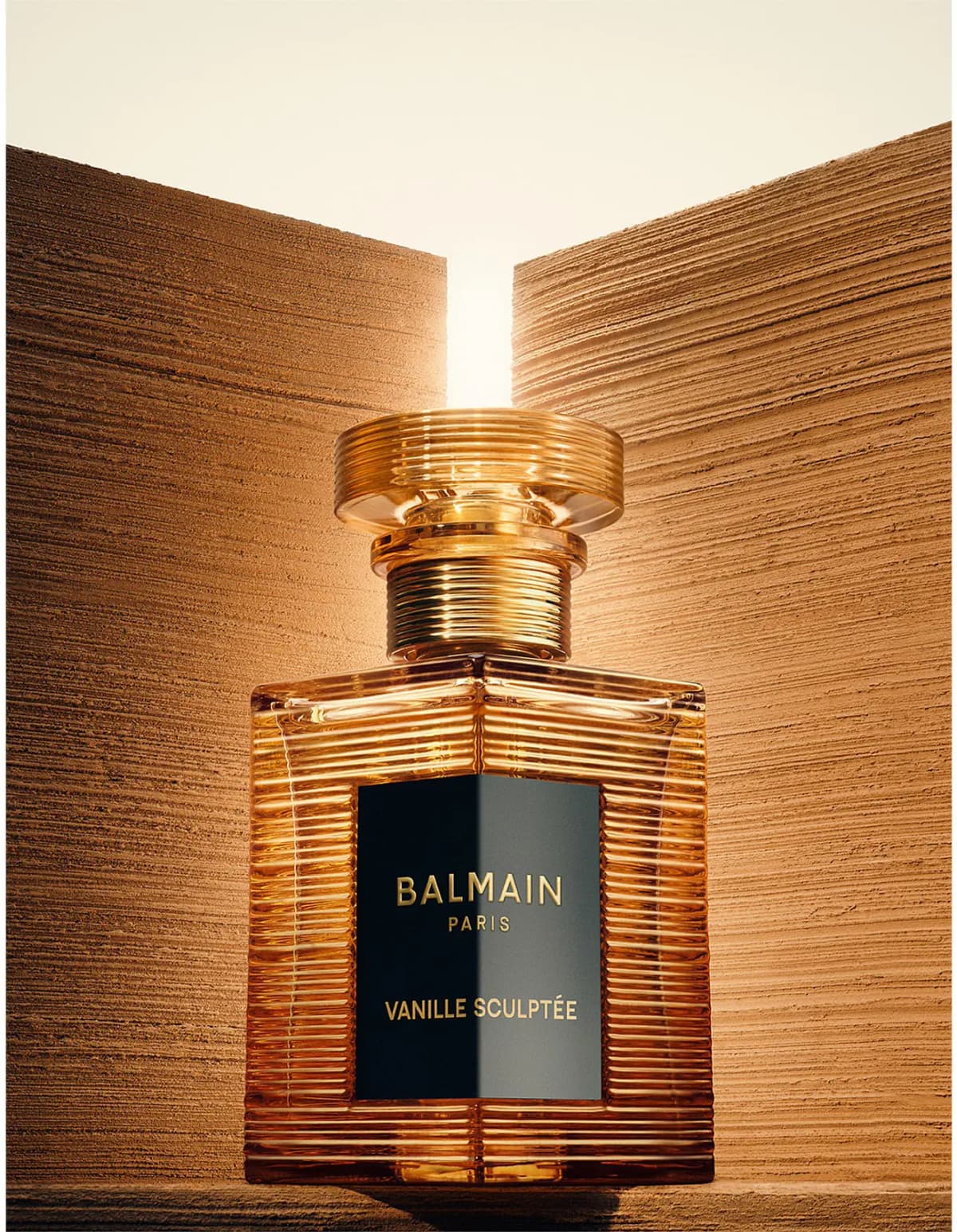Balmain Vanille Sculptée ribbed amber-gold bottle glowing between textured sandstone walls in warm light