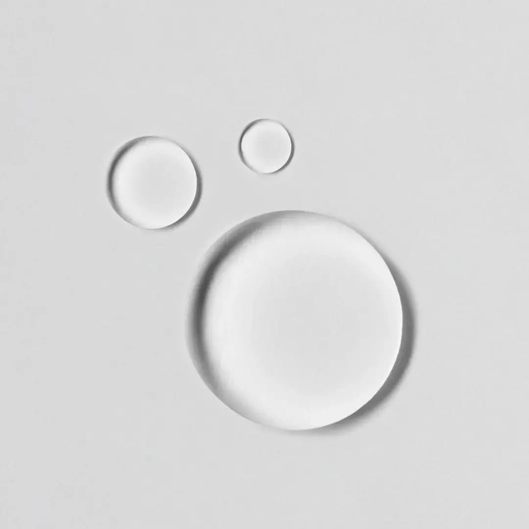 Three clear glossy droplets of liquid in varying sizes resting on a pale grey surface