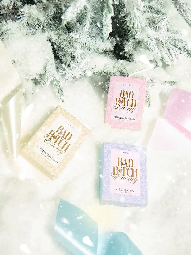 Flat lay of three P Louise Bad Bitch Energy packs in pink, blue and cream on snow with frosted pine branches.