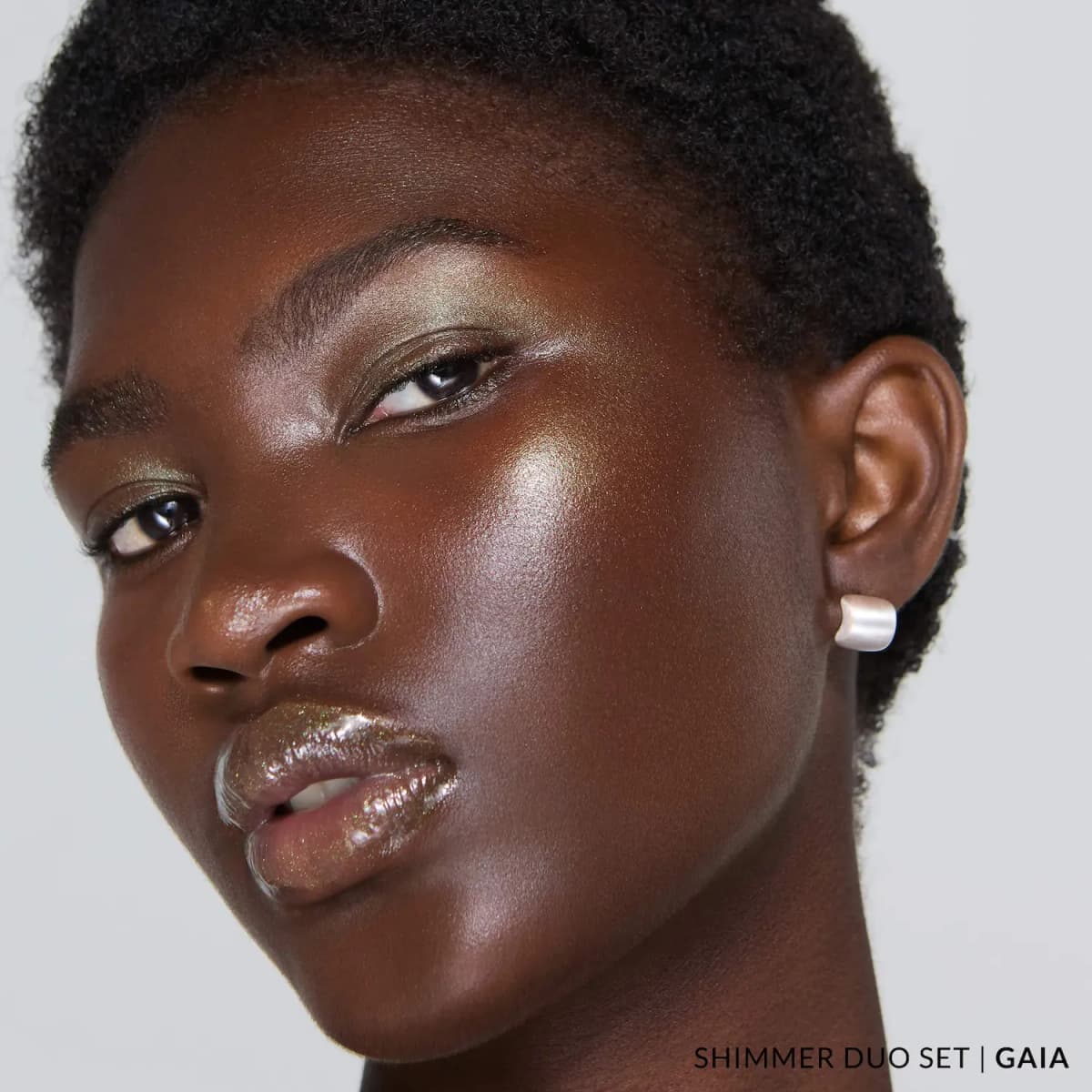 Model with short natural hair wearing Gaia: green-gold shimmer on eyes and cheeks and glittery clear gloss on lips