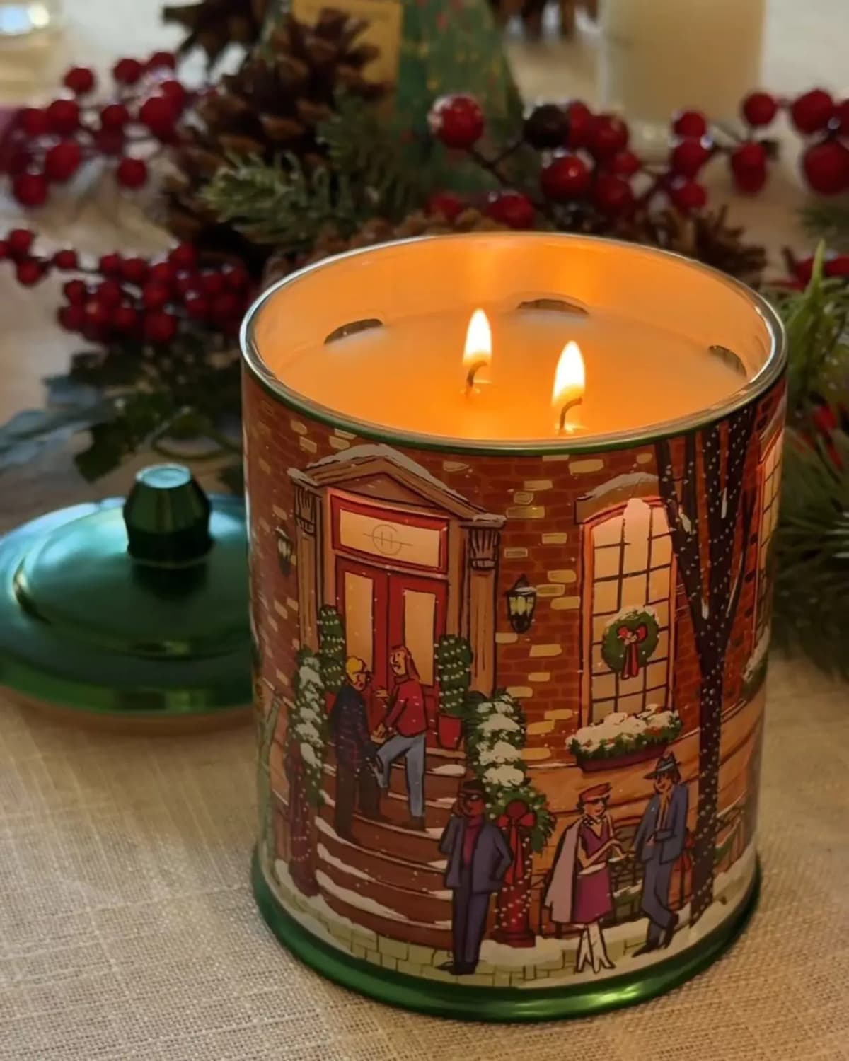 Lit brownstone candle with two flames and glowing windows beside its green lid, with red berries and pine cones behind