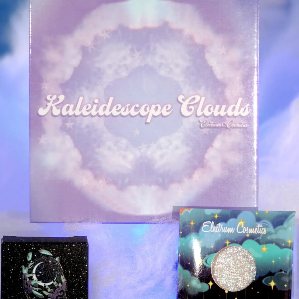 Electrum Cosmetics Kaleidoscope Clouds eyeshadow palette box beside a black moon-print box and a glittery single pan