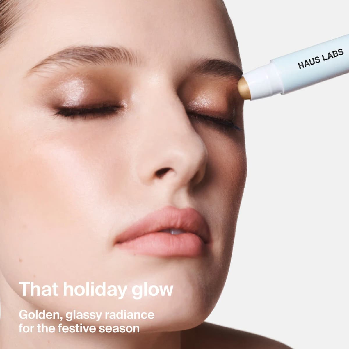 Model with eyes closed as the gold balm stick is dabbed on the inner eye, with "That holiday glow" text