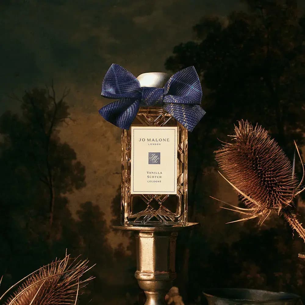 Cut-glass cologne bottle with blue tartan bow on a brass candlestick beside dried teasels against a dark landscape painting