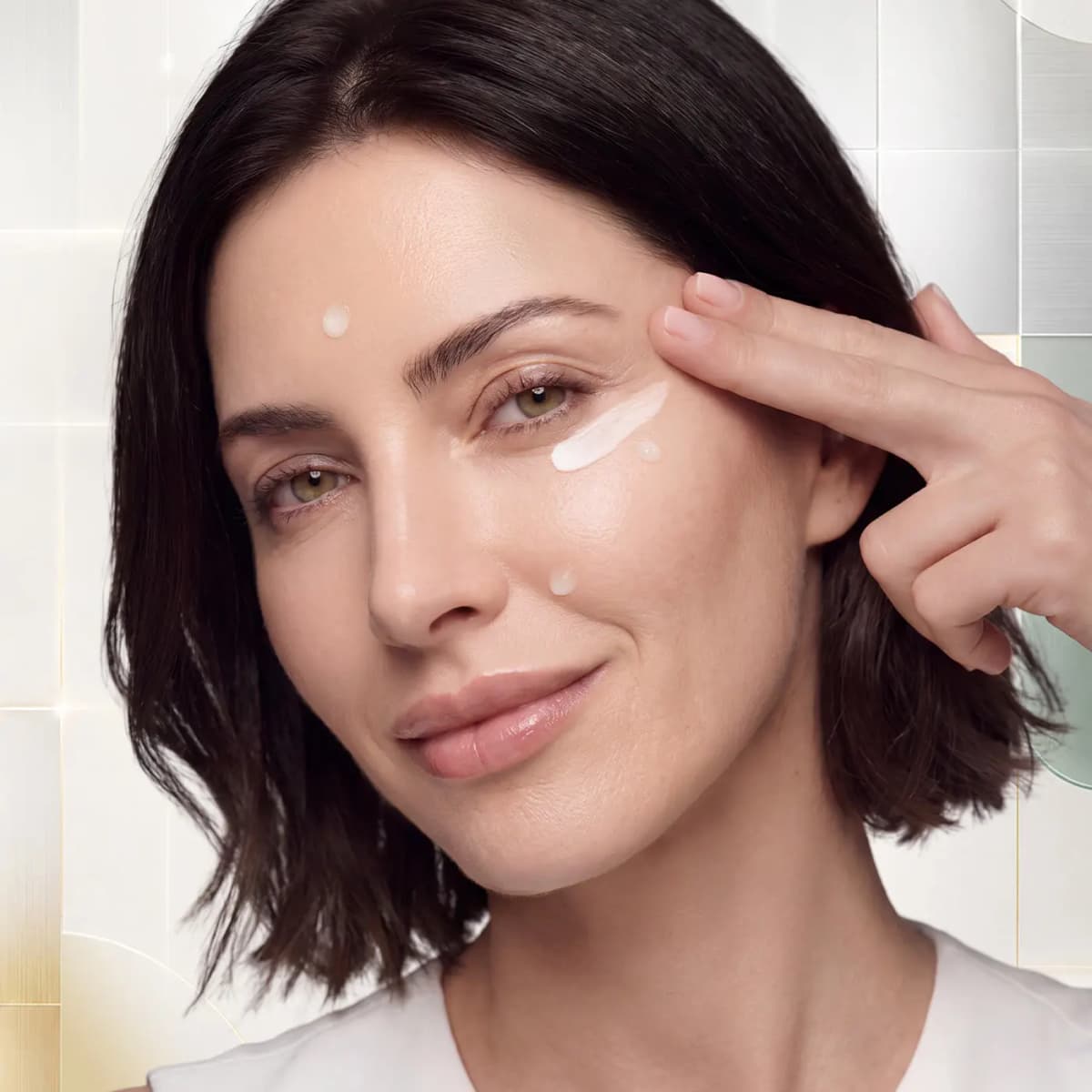 Dark-haired model smiling as she pats a white cream streak under her eye, with dots of cream on her face