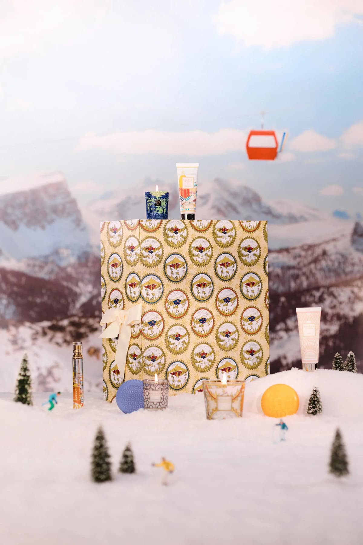 Wrapped calendar with cream bow standing in a miniature snow scene with candles, hand cream, soaps and a red gondola overhead
