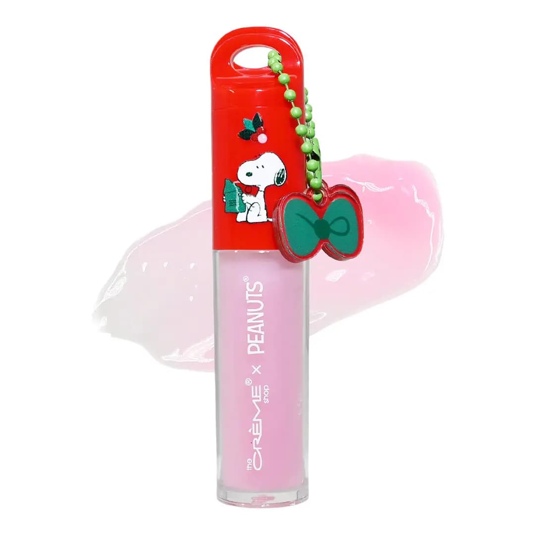 Pink Afterglow lip oil with red Snoopy cap and green bow charm on a beaded chain, over a sheer pink swatch.