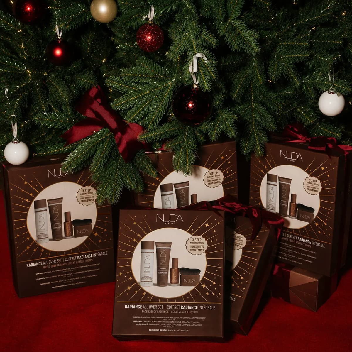 Several NUDA Radiance All Over Set boxes with red velvet bows beneath a decorated Christmas tree on red carpet.