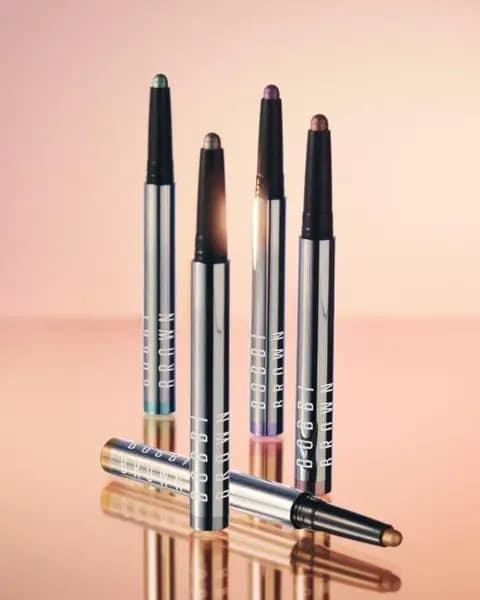 Five Bobbi Brown shadow sticks in silver chrome tubes, one lying down with a gold tip, on a reflective peach surface.