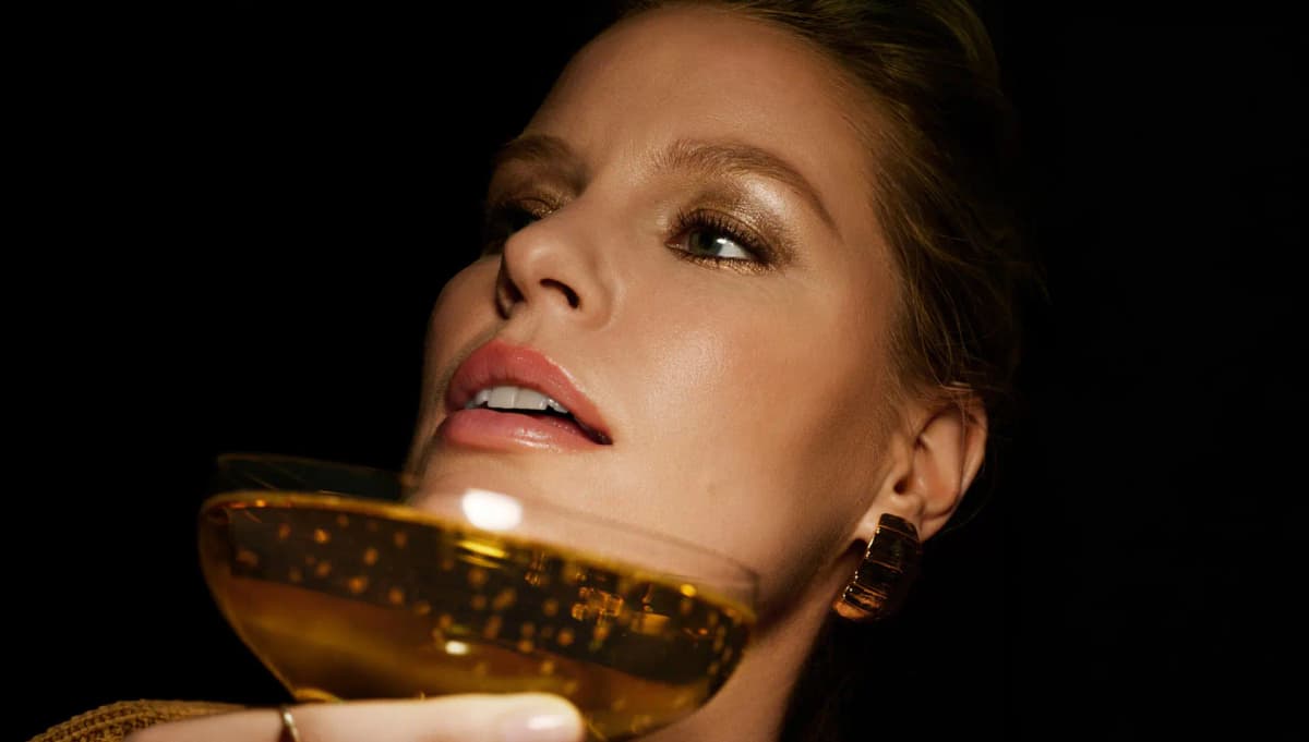 Woman with glowing bronze eye makeup holding a coupe of golden champagne against a black backdrop.