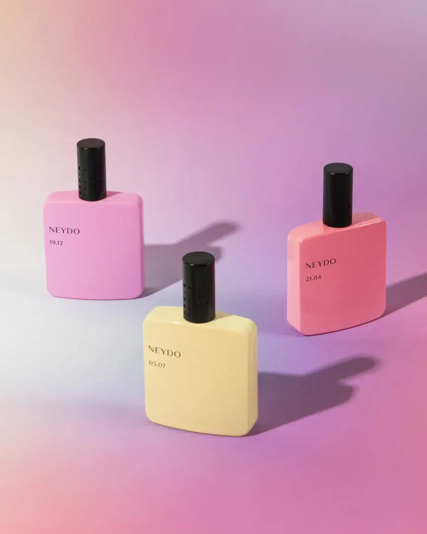 NEYDO 19.12, 05.07 and 21.04 bottles in lilac, pale yellow and pink with black caps on a pink-purple gradient backdrop