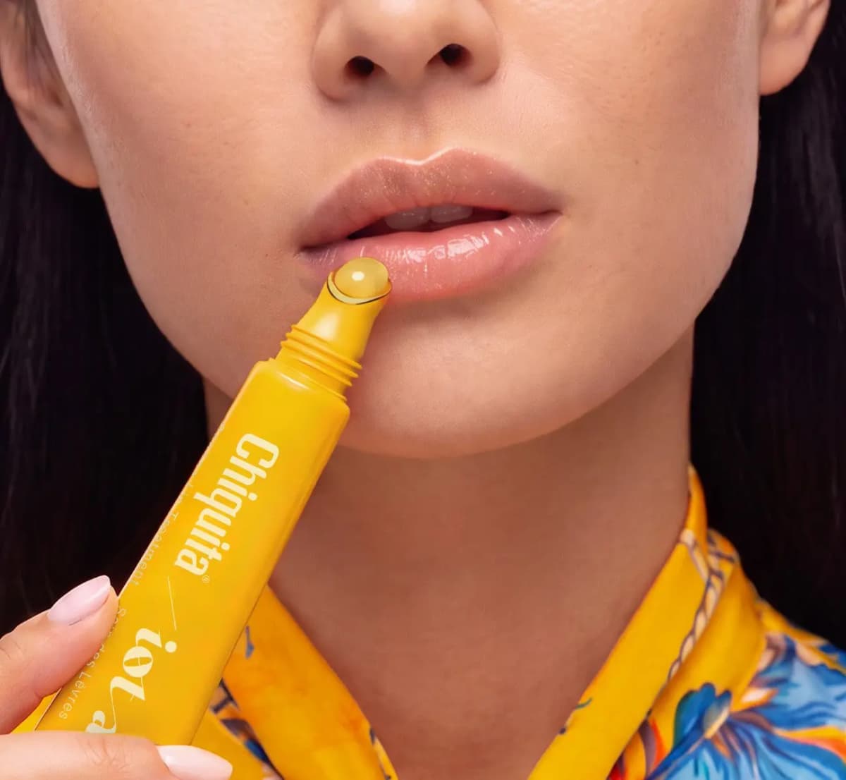 Model in a floral yellow shirt applying the clear treatment, leaving lips glossy and natural pink