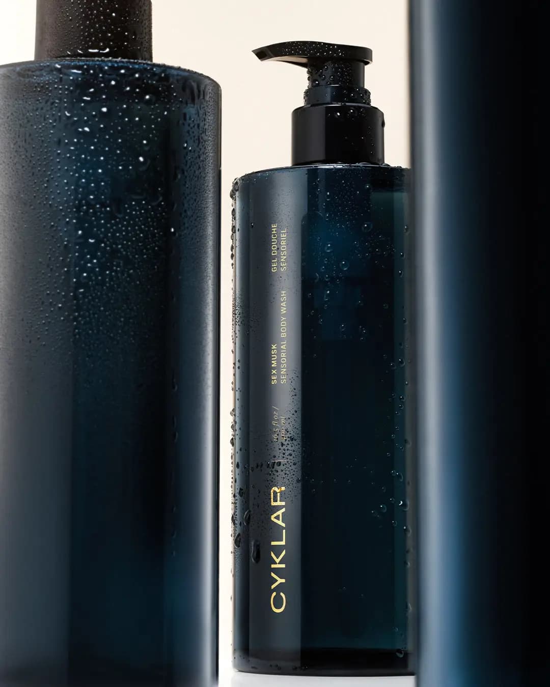 Water-beaded navy body wash bottle with gold label text, framed between two blurred dark bottles