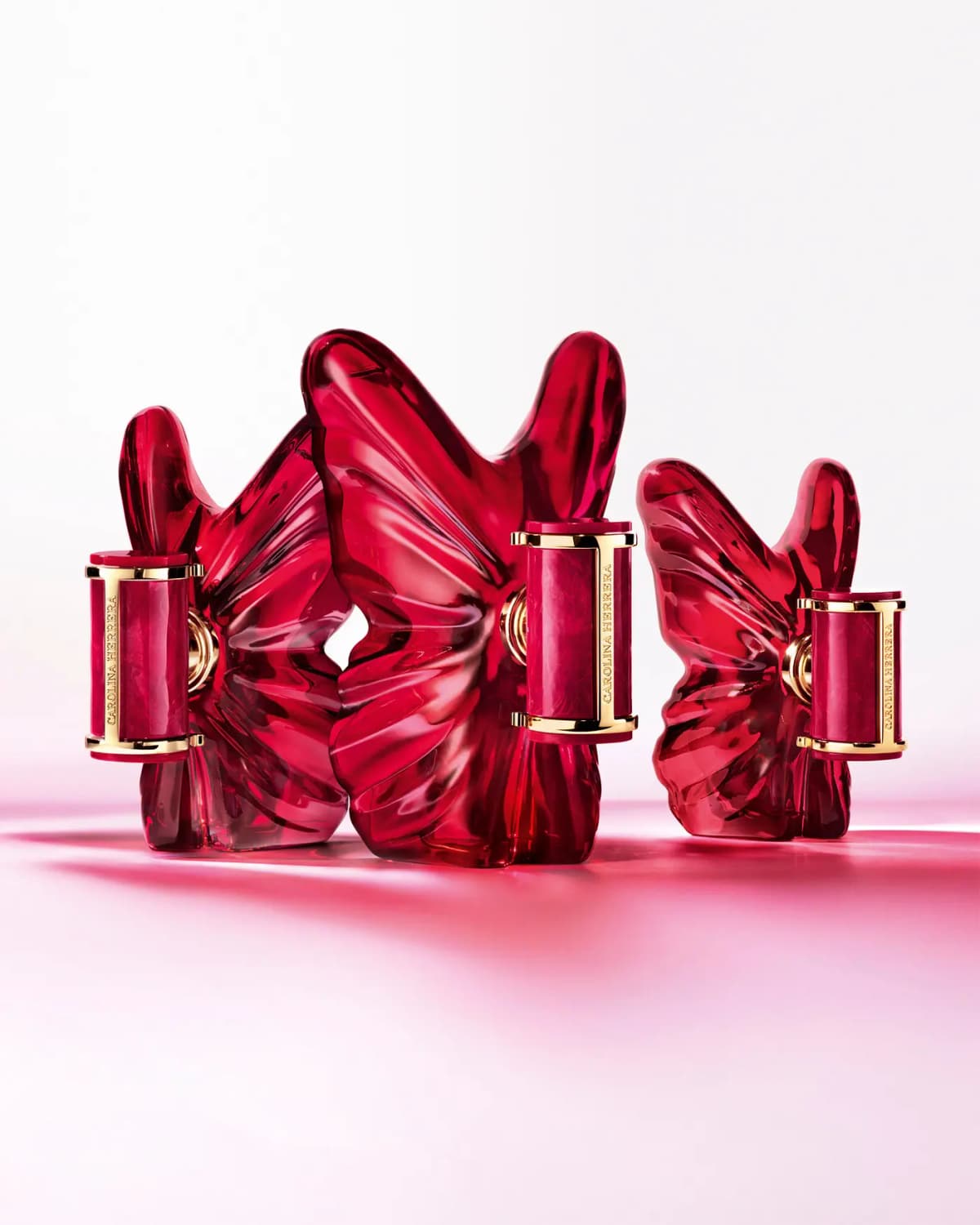 Three red butterfly bottles of different sizes with gold-trimmed marbled caps on a pink reflective surface