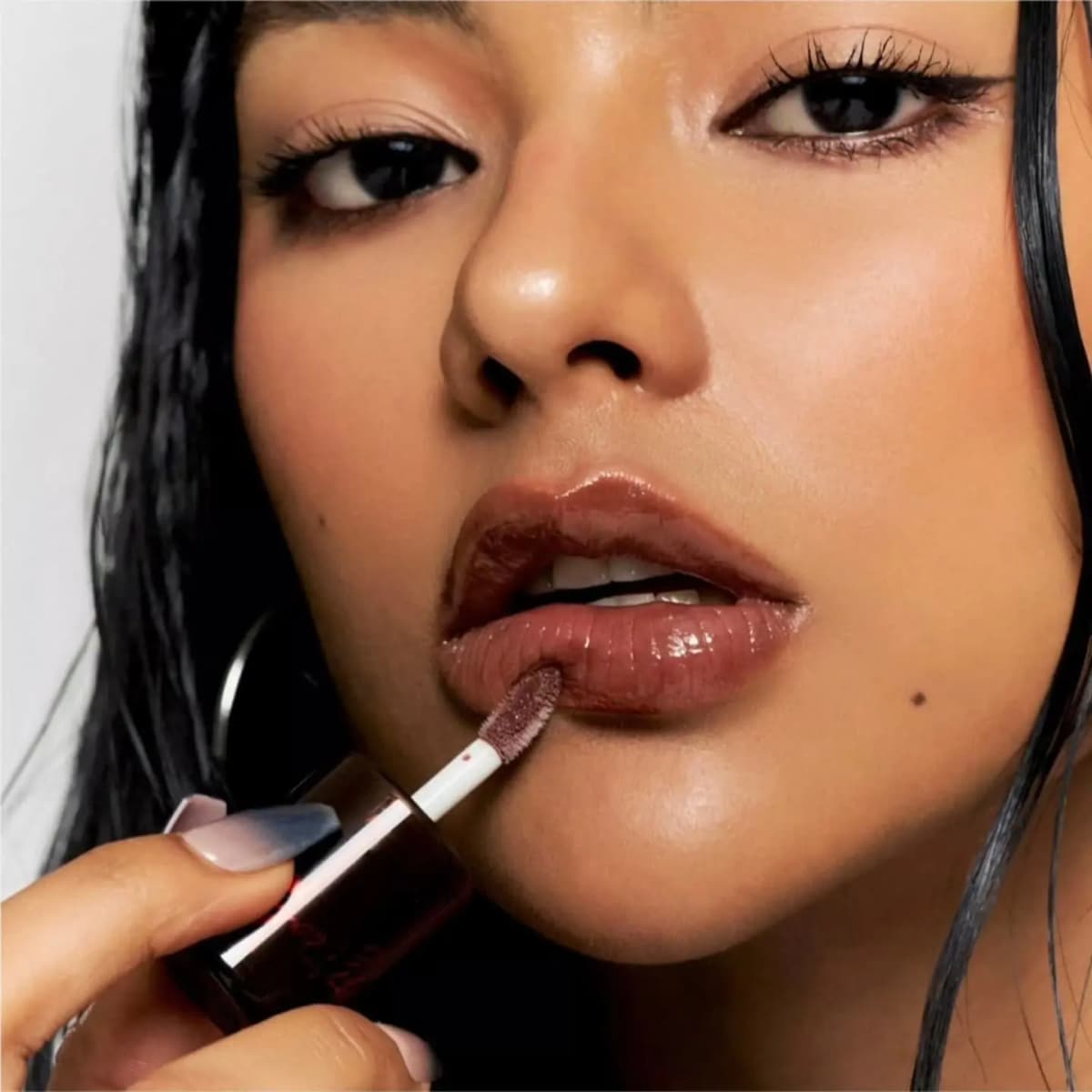 Model with long black hair applying glossy brown gloss with a doe-foot wand from a dark amber bottle.