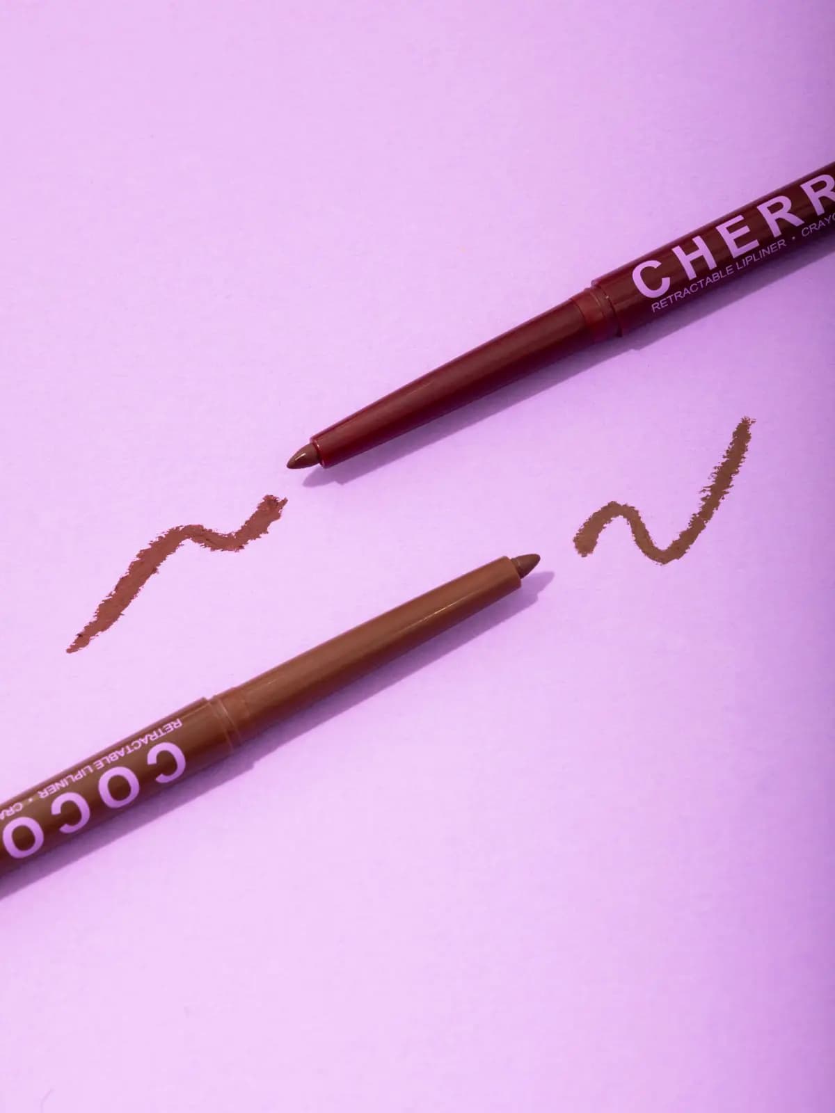 Two retractable lip liners, burgundy Cherry and brown Cocoa, with matching squiggle swatches on lilac paper