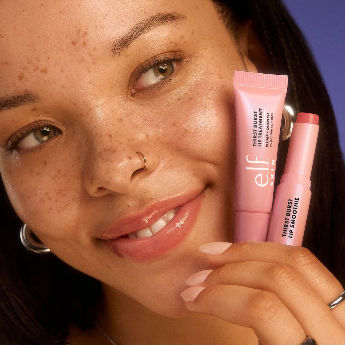 Smiling freckled model with glossy lips holds the Thirst Burst Lip Treatment and Lip Smoothie by her cheek.