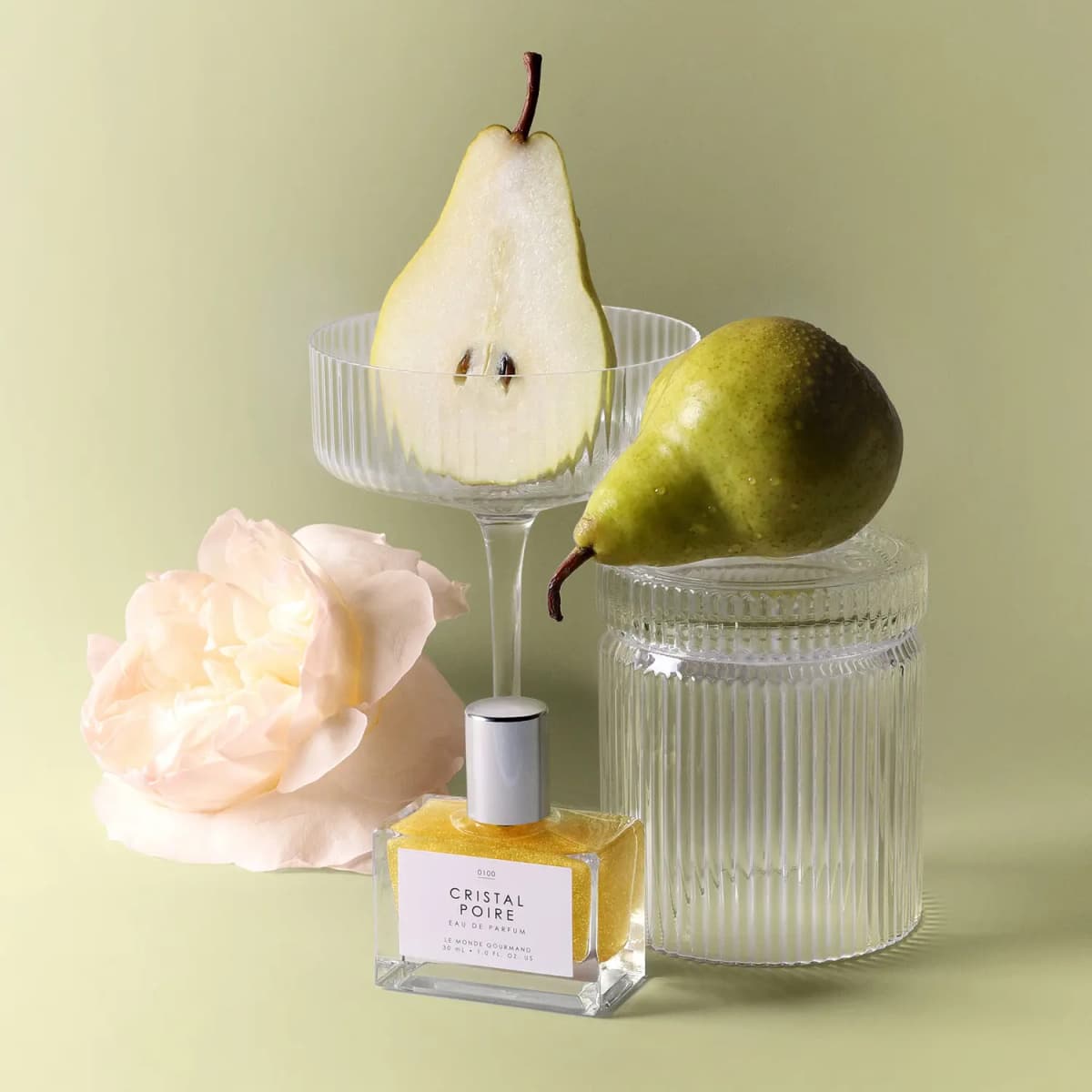 Shimmering gold perfume beside a pale rose, halved pear in a ribbed coupe glass and whole pear on a glass jar
