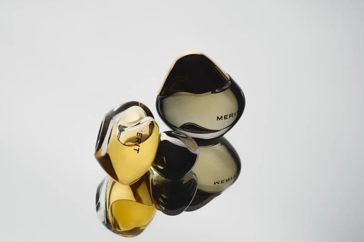 Golden bottle and chrome cap lying beside an upright smoky dark-tinted MERIT bottle on a mirrored surface