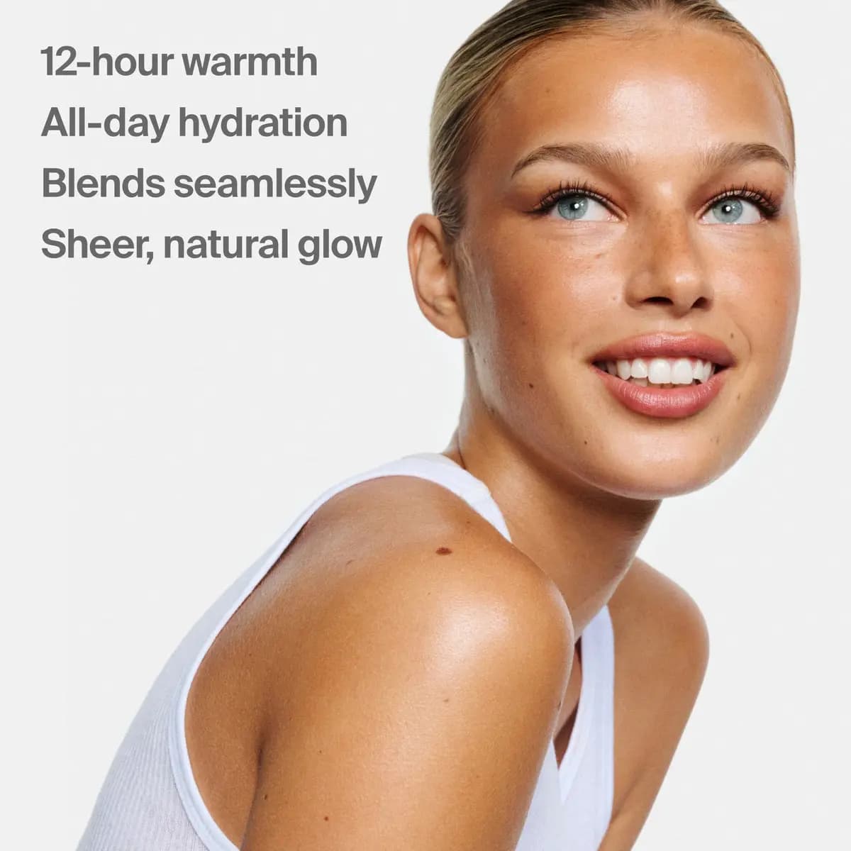 Smiling freckled blonde model with glowing bronzed skin in a white tank, with text '12-hour warmth' and 'Sheer, natural glow