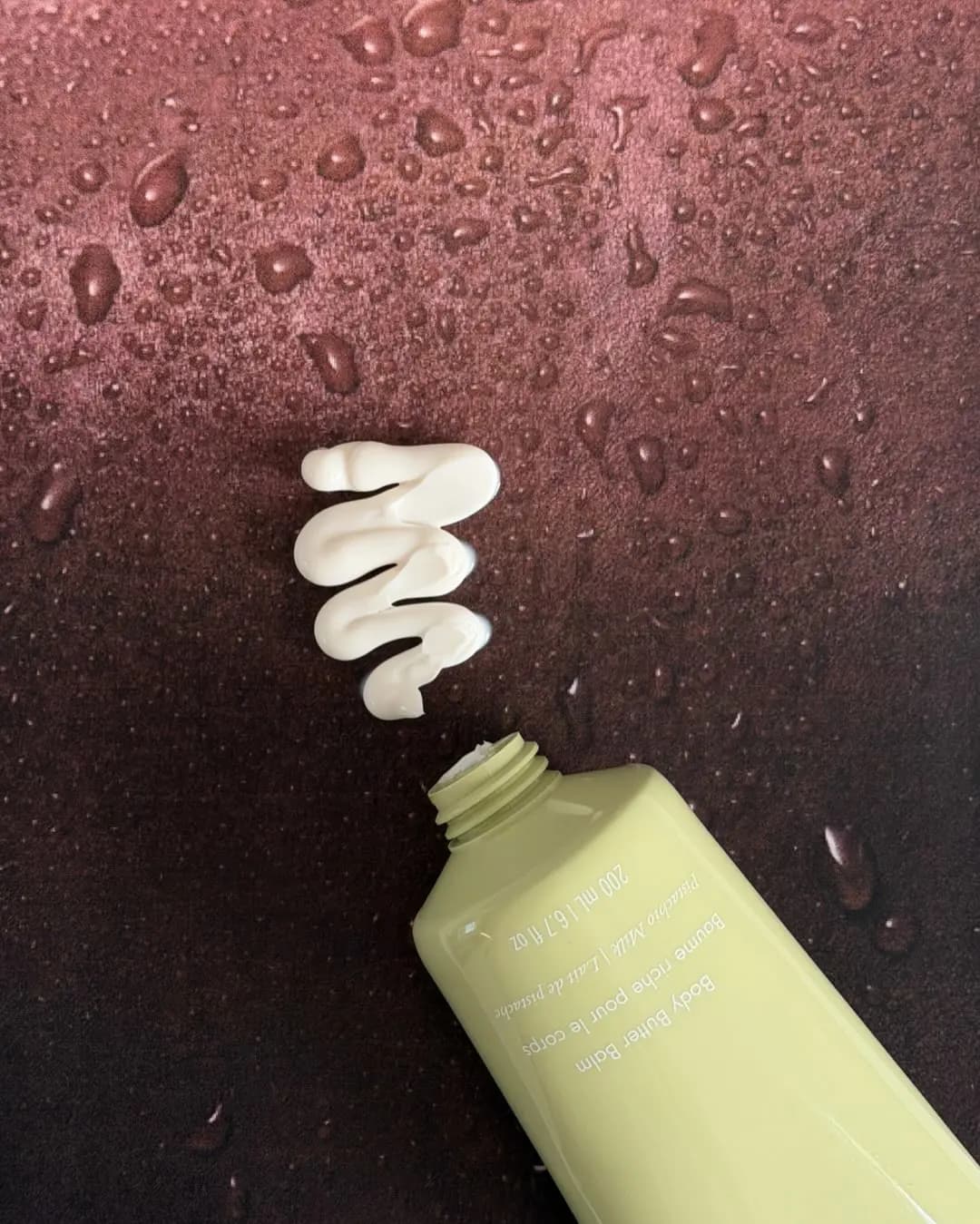 Open green Body Butter Balm tube beside a squiggle of ivory cream on a burgundy surface dotted with water droplets.